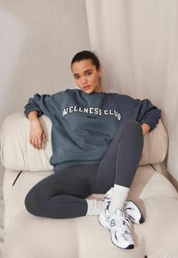 Šedá mikina s nápisom "WELLNESS CLUB", kombinovaná s tmavými legínami a bielymi športovými topánkami, sedí na svetlofarebnej čalúnenej stoličke.