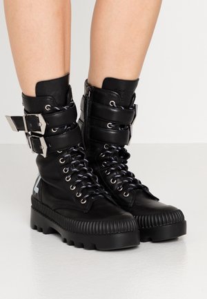 Schwarze Leder-Kampfstiefel mit dicken Sohlen, Schnürsenkeln, seitlichem Reißverschluss, doppelten Riemen mit silbernen Schnallen, getragen an bloßen Beinen.