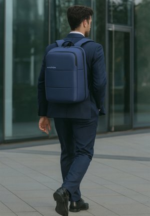 Blaue Tasche mit glatter Oberfläche, rechteckiger Form, schwarzem Reißverschluss und Logo. Trägt eine Person im Anzug, die auf einem gefliesten Boden geht.