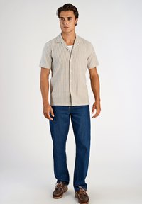 Jasnobeżowa koszula z krótkim rękawem w pionowe paski, zapinana na guziki. Połączona z ciemnoniebieskimi jeansami i brązowymi mokasynami.