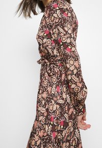 Abito floreale marrone con maniche lunghe, caratterizzato da una vita aderente e una cintura. Il tessuto presenta intricati motivi di fiori e foglie.