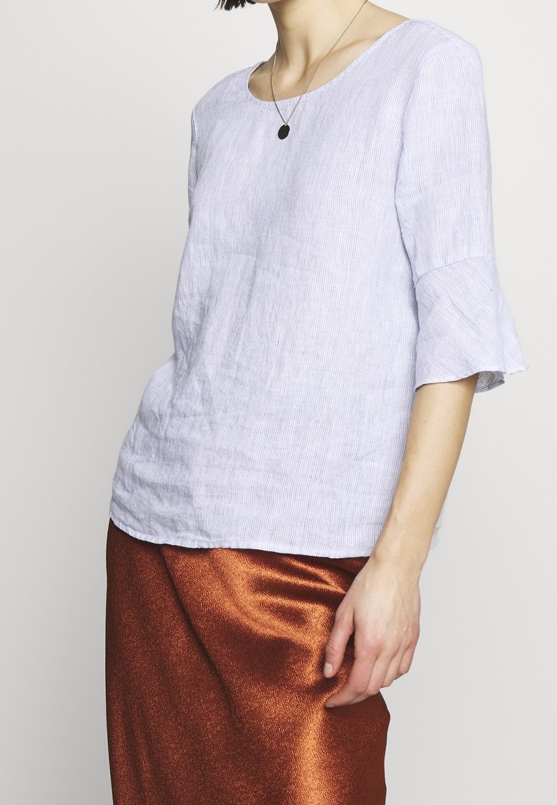 Top in lino a righe azzurro chiaro con maniche a campana, scollatura rotonda, abbinato a una gonna bronzo lucido. Focus sulla texture e sul contrasto di colori.