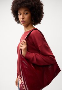 Borsa tote in vernice bordeaux con texture liscia, tenuta da una persona che indossa un maglione abbinato. Stile casual, forma oversize, design minimal.