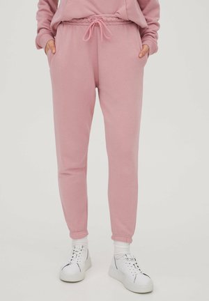 Personne portant un pantalon de survêtement rose à cordon, mains dans les poches et baskets blanches avec chaussettes blanches sur fond uni.