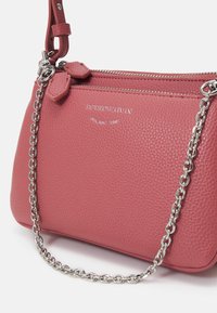 Růžová texturovaná kožená kabelka se stříbrným řetízkovým popruhem a zipem, s vyraženým logem značky a dekorativními přívěsky na zipu.