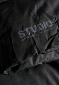 Tuviena melnās auduma tuvplānā ar uzrakstu "STUDIO" lielām burtiem un mazāku tekstu "JXX® IZVEIDOTS DIVDESMIT DIVDESMIT".