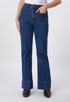 Moteris dėvi mėlynus džinsus su aukštu liemeniu, priekinėmis kišenėmis, matomomis vertikaliomis siūlėmis ir rudais aulinukais su kvadrato formos pirštais.