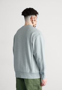 Sudadera gris claro con cuello redondo acanalado, mangas largas y corte relajado. La tela parece suave, con costuras visibles y una textura lisa.
