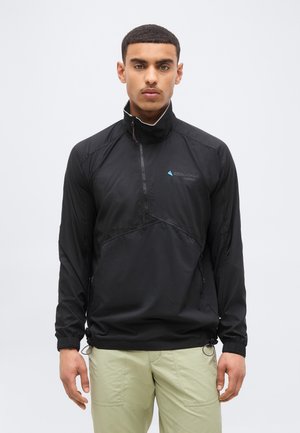 Man draagt een zwarte lichte pulloverjas met kwartrits en beige broek, staand tegen een effen witte achtergrond.