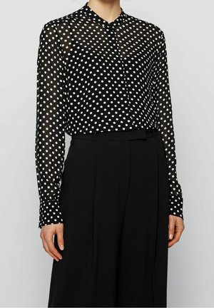 Mujer con una blusa negra transparente de manga larga con lunares blancos metida dentro de unos pantalones negros de talle alto.