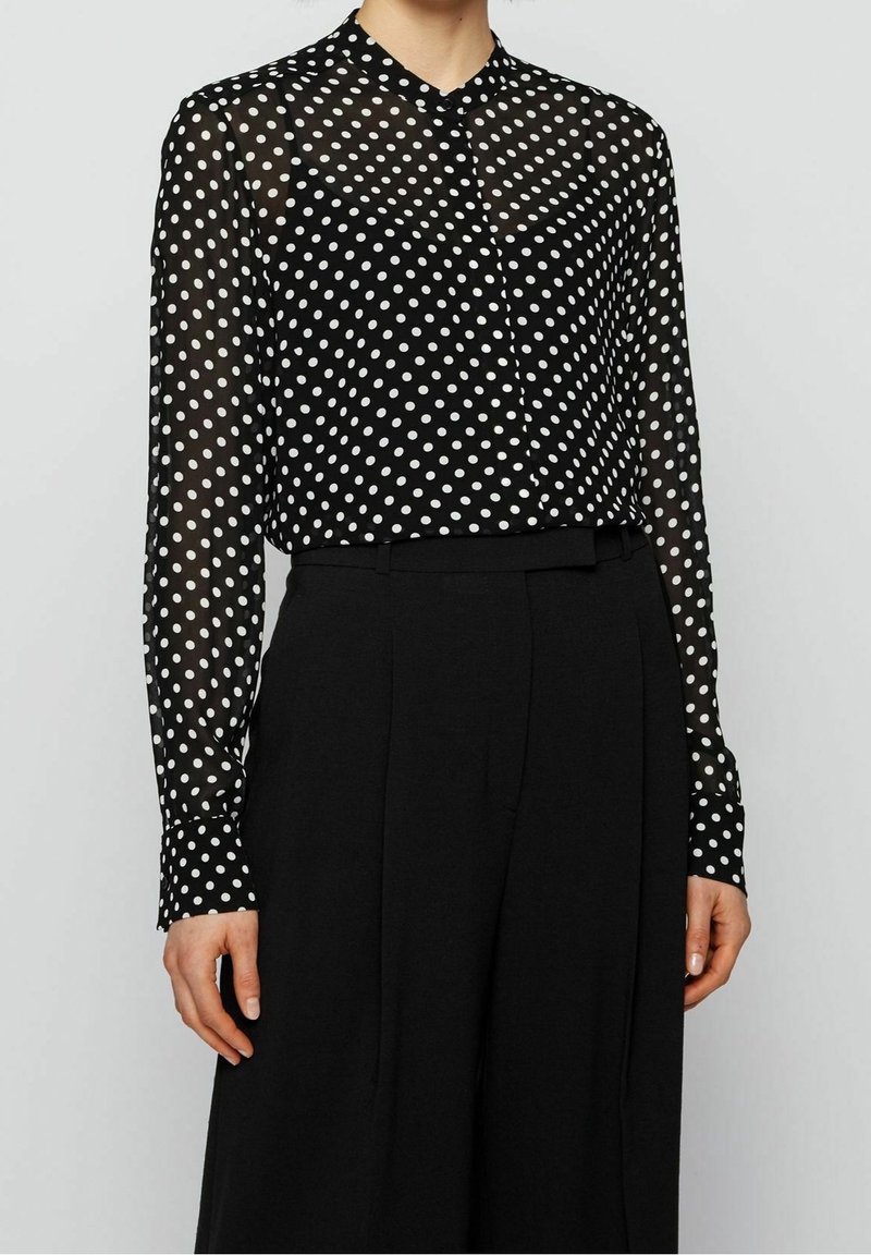 Femme portant un chemisier noir transparent à manches longues avec des pois blancs, rentré dans un pantalon noir taille haute.