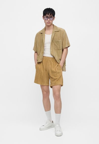 Jeune homme portant une chemise à manches courtes couleur sable à boutons et un short assorti, un débardeur blanc, des chaussettes blanches, des baskets blanches et de grandes lunettes teintées de violet.