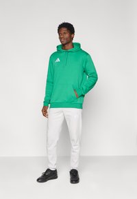 Πράσινο φούτερ με κουκούλα, μπροστινή τσέπη και κορδόνι, σε συνδυασμό με λευκή φόρμα με λογότυπο. Μαύρα αθλητικά παπούτσια ολοκληρώνουν το σύνολο.