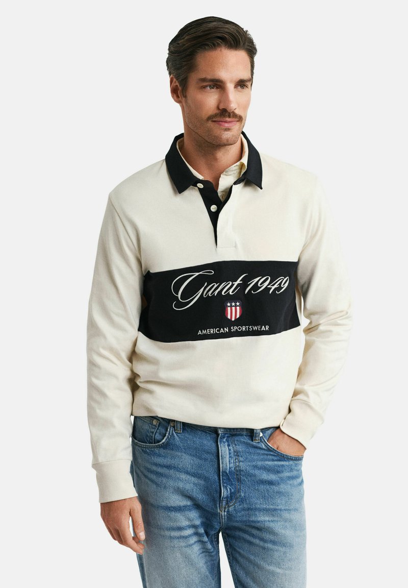 Polo blanc à manches longues avec col noir et bande horizontale noire affichant le logo "Gant 1949". Porté avec un jean bleu clair.