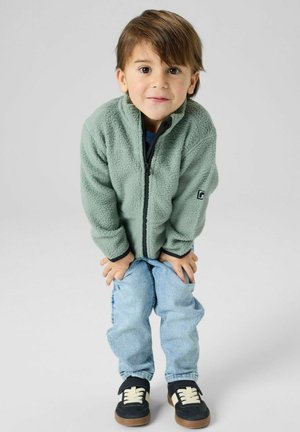 Jeune enfant portant une veste polaire verte, un jean bleu clair et des baskets noires, debout avec les mains sur les genoux devant un fond uni.