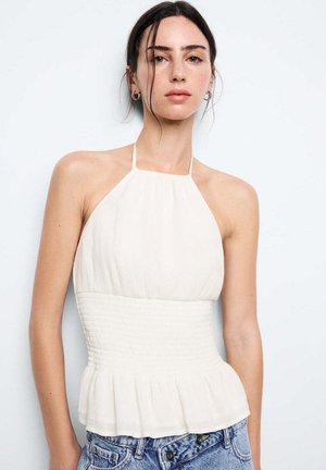 Jeune femme aux cheveux foncés porte un haut blanc sans manches à col halter et un jean bleu sur un fond clair uni.