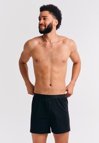 Bovenlichaamloze man met een baard en krullend haar, gekleed in een zwarte boxershort, die zijn tailleband aanpast terwijl hij naar de zijkant kijkt tegen een effen achtergrond.
