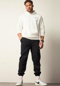 Sudadera blanca con capucha, bolsillo frontal y logo negro, combinada con joggers negros ajustados y zapatillas blancas con detalles en negro.