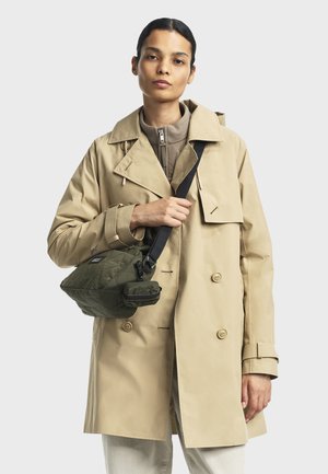 Kvinna i beige trench coat och vadderad jacka, som håller en grön vadderad crossbody-väska med svart rem mot en enfärgad bakgrund.