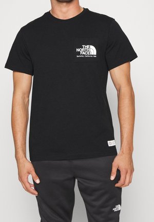 Mężczyzna w czarnej koszulce The North Face i dopasowanych czarnych spodniach z logo marki na klatce piersiowej i udzie na jednolitym tle.