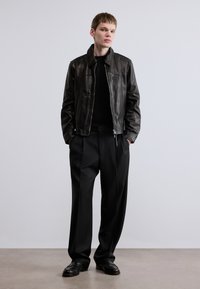 Μαύρο δερμάτινο μπουφάν με όρθιο γιακά, σε συνδυασμό με μαύρο πουλόβερ και ραμμένο μαύρο παντελόνι. Το μοντέλο στέκεται με τα χέρια στις τσέπες.