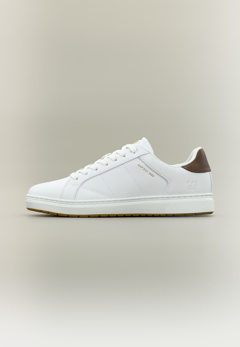 Baskets basses blanches avec languette de talon marron, lacets blancs, et texte "GAP EST. 1969" en or sur le côté.
