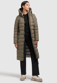 Long olive green puffer coat with a hood, featuring horizontal quilted design, front zipper, and side slits, paired with black pants and chunky footwear.