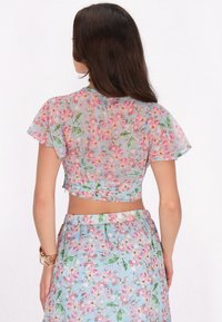 Bauchfreies Top aus transparenter Stoff mit pastellfarbenen Blumenmustern in Pink und Grün auf einem hellblauen Hintergrund; mit schwingenden Ärmeln und taillierter Passform.