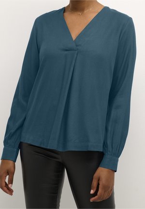 Femme portant un chemisier bleu sarcelle à manches longues avec col en V et poignets boutonnés, ainsi qu’un pantalon noir ajusté, visible des épaules jusqu’aux hanches.