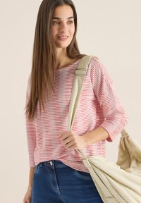 Kvinde med langt brunt hår iført en trøje med røde og hvide striber og blå jeans, bærende en beige taske over skulderen.