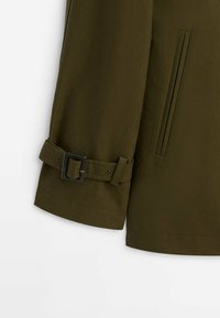 Veste vert olive avec une texture lisse, dotée d'un poignet ceinturé, de deux poches latérales et d'une coupe droite. Détail de couture subtil.
