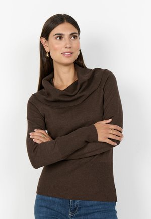Kvinna med långt mörkt hår som bär en brun tröja med krage och blå jeans, med armarna korsade och tittande lite åt sidan, neutral bakgrund.