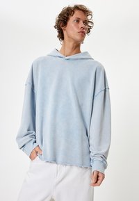 Sudadera azul claro de gran tamaño con una textura suave, hombros caídos y dobladillo sin terminar. Presenta una capucha y puños de manga acanalados para mayor comodidad.