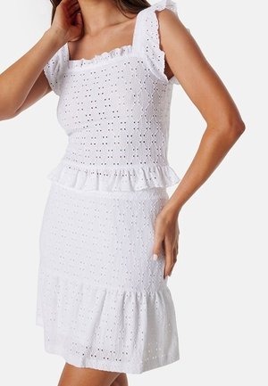 Femme portant une robe blanche sans manches en œillets avec un col carré volanté, à la taille et à l'ourlet, posant une main près du cou et l'autre sur la hanche.