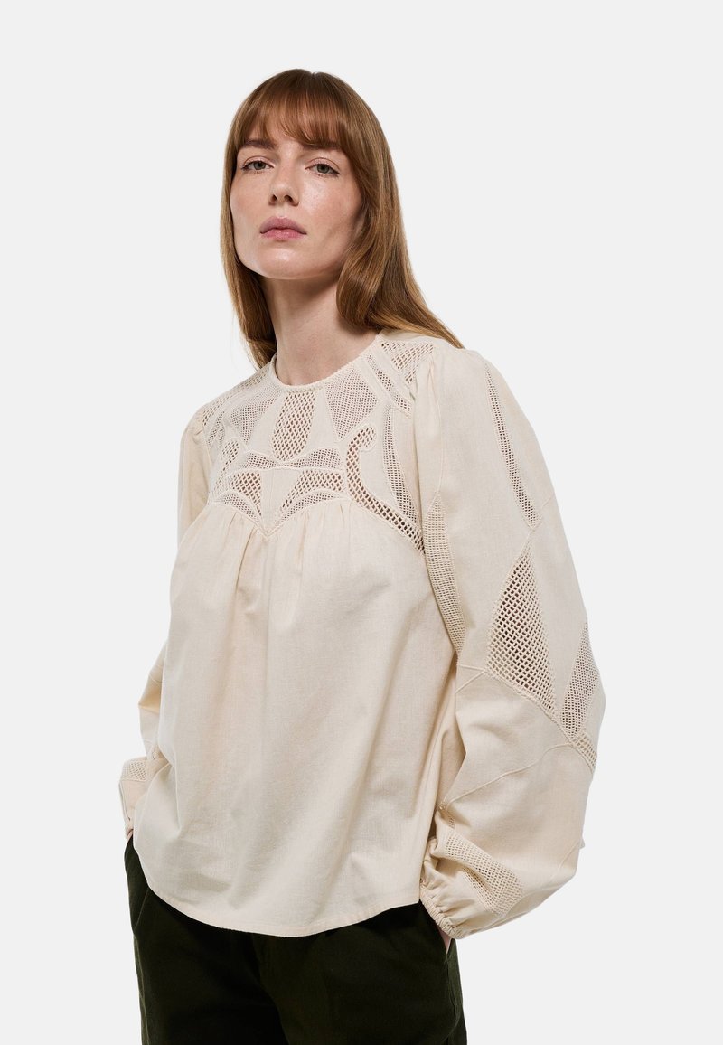 Blouse de couleur crème avec un empiècement en dentelle texturée et des manches ballons avec un ourlet froncé, confectionnée dans un tissu léger.