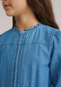 Kék farmerruházat elöl cipzárral és fodros gallérral. Gyűrt vállakkal és sima textúrával rendelkezik. Hétköznapi kialakítás.