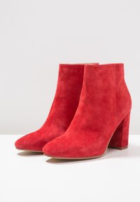 Bottes chevilles en daim rouge avec une texture lisse, bout pointu et talon. Le design est élégant avec des lignes épurées et peu de détails.