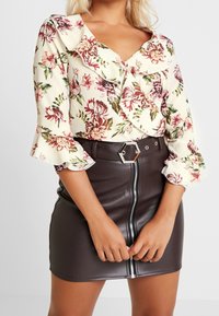 Blusa floreale con colletto e polsini arricciati, sfondo crema, motivi rossi e verdi; abbinata a una minigonna di pelle marrone aderente con cerniera frontale.