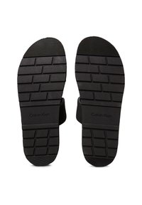Schwarze Sandalensohlen mit einer strukturierten Grip-Oberfläche. Enthält ein "Calvin Klein"-Logo. Glattes Gummimaterial mit unterschiedlichen Profilmustern.