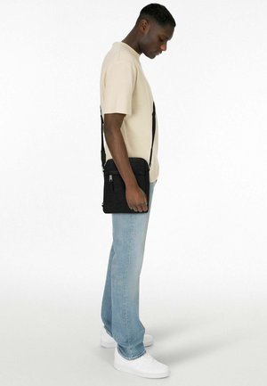 Sac bandoulière noir à surface texturée, poche zippée à l'avant et sangle réglable, porté par un homme en chemise beige et jean clair.