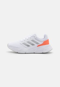 Zapatilla deportiva blanca con upper de malla, tres franjas grises en el costado, suela blanca y detalle en el talón de color naranja brillante, mostrada de perfil sobre un fondo blanco.