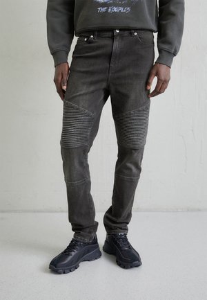 Homme portant un jean noir côtelé avec un design au niveau des genoux et des baskets noires épaisses, debout contre un mur uni.