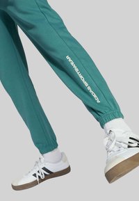 Tyrkysové tepláky s elastickými manžetami, so bielym logom "Adidas Sportswear" pozdĺž strany. Kombinované s bielymi teniskami a krátkymi ponožkami.