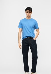 Νεαρός άνδρας που φοράει καφέ γυαλιά ηλίου, μπλε μπλουζάκι, σκούρο τζιν με μαύρη ζώνη και λευκά αθλητικά παπούτσια, στέκεται μπροστά σε απλό φόντο.