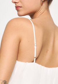 Vit tygklänning med tunna, justerbara axelband. Har en mjuk, texturerad yta och en rundad halsringning som visar naken hud på ryggen.