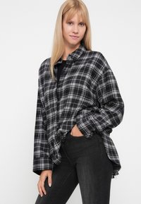 Cămașă din flanel cu carouri negre și gri, cu o croială lejeră, guler butonat și umeri căzuți. Purtați peste un top negru și asortată cu blugi închiși.
