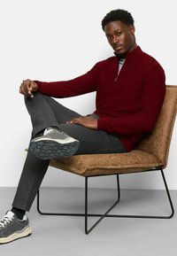 Burgunderfarbener Reißverschluss-Pullover mit einem strukturierten Muster, kombiniert mit dunklen Jeans und grauen Sneakers, auf einem braunen gepolsterten Stuhl sitzend.