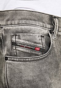 Harmaat farkut, joissa on takatasku, jossa on merkkilappu. Kangas on hieman kulunut ja ompeleissa on kontrastiväriä.