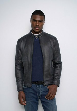 Joven con chaqueta de cuero negra, suéter azul marino y vaqueros azules, de pie frente a un fondo claro y sencillo.