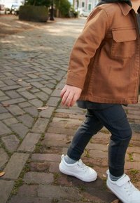 Beige zip-up jack met twee zakken, gedragen over een grijze shirt, gecombineerd met donkergrijze broek en witte sneakers, wandelend op een stenen pad.